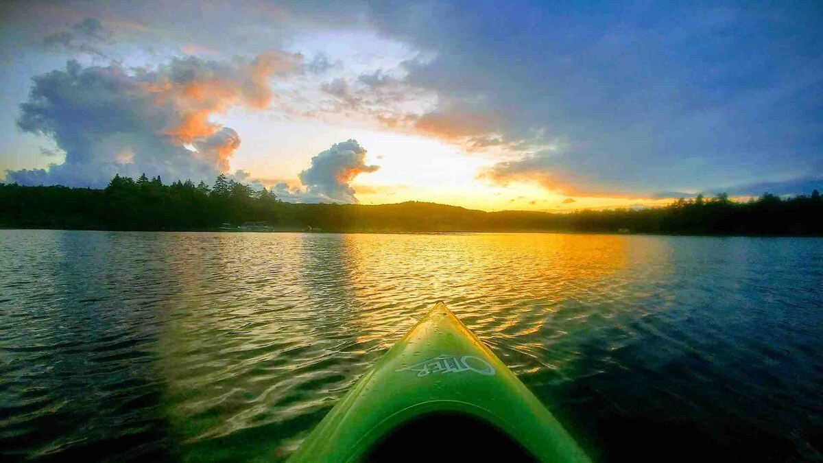 A kayak is seen cutting through the calm water, framed by a vibrant sunset reflecting warm hues across the lake. The sky is adorned with clouds, adding depth to the tranquil scene. Lush trees silhouette the horizon, completing the serene natural landscape.