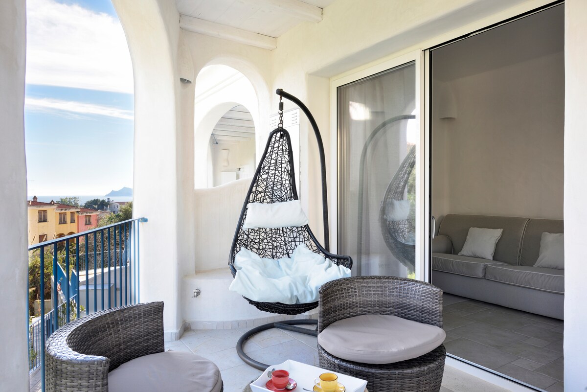 A serene outdoor terrace features a hanging chair with soft cushions, accompanied by two plush wicker chairs. A coffee table displays cups, enhancing the inviting space. Large sliding glass doors provide access to an airy living area, with scenic views of the surroundings.