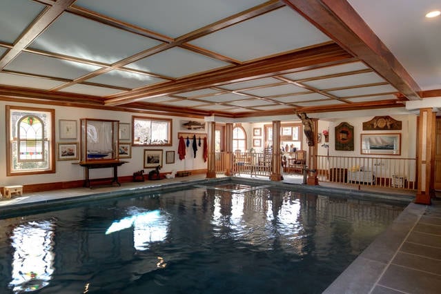 An indoor heated pool is surrounded by elegant wood paneling and framed artwork. Natural light filters through the windows, reflecting off the water's surface. A cozy sitting area is positioned nearby, offering a relaxing space adjacent to the pool.