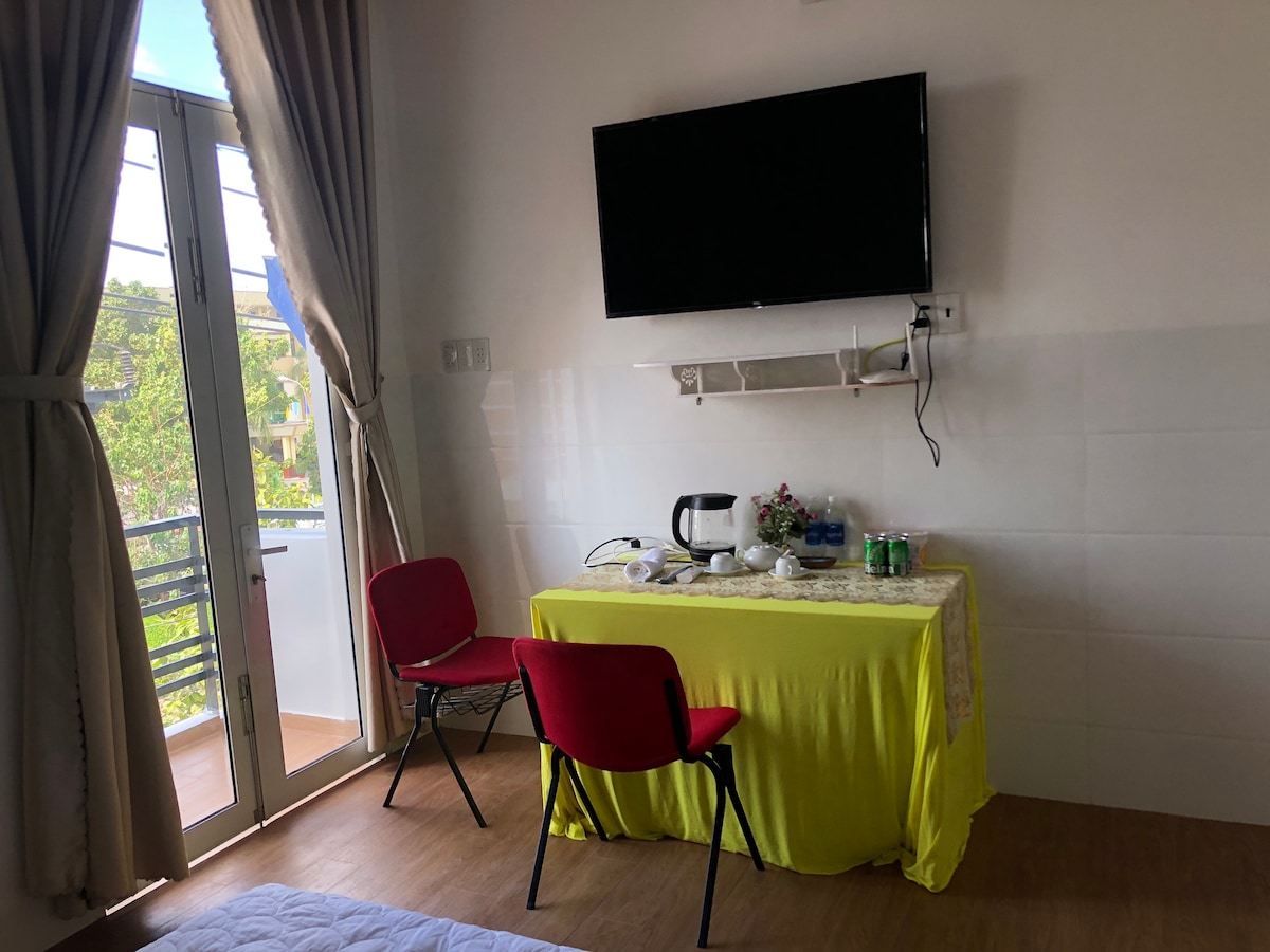 A compact corner of the room features a small table covered with a bright yellow cloth. A kettle and several beverage cans are positioned on the table, accompanied by a red chair. Large windows are draped with light-colored curtains, allowing natural light to enter.