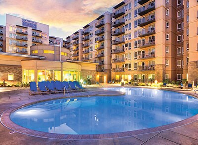 An outdoor pool area is surrounded by a spacious deck featuring lounge chairs. The multi-story building with balconies provides a backdrop, while soft evening light enhances the peaceful environment. Plants are placed around the pool for a touch of greenery.