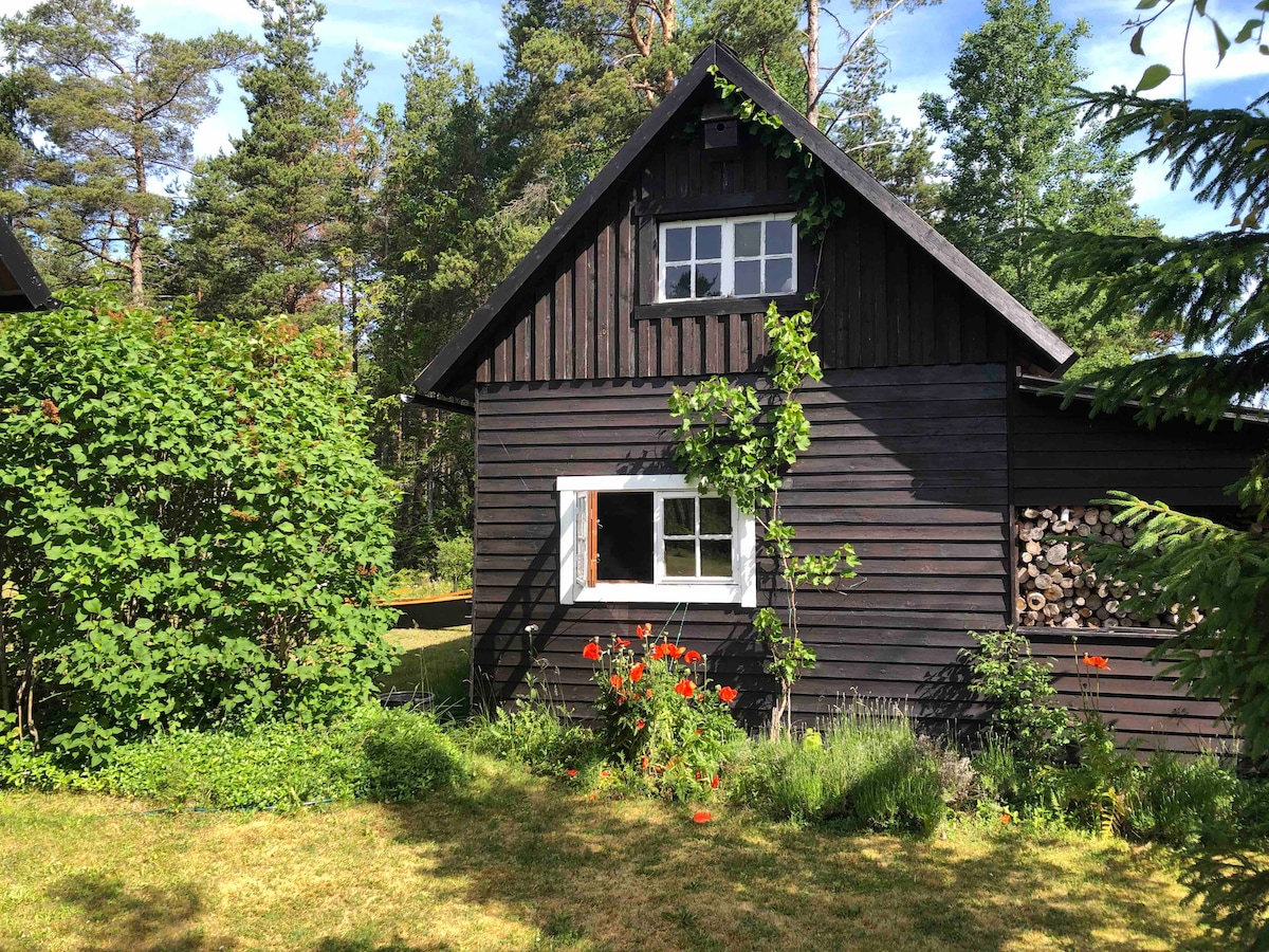 A charming wooden guesthouse is nestled among lush greenery, with a black façade and white window accents. Wood logs neatly stacked beside the building enhance the rustic appeal. Bright flowers add a splash of color to the garden, creating a welcoming outdoor environment.