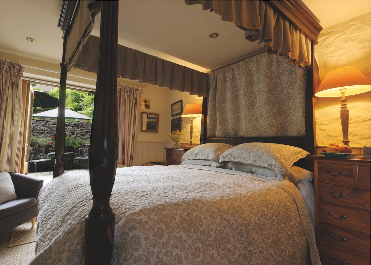 A four poster bed is positioned in a softly lit room, adorned with elegant bedding. Natural light enters through open doors leading to a private courtyard. Two bedside tables with lamps are placed on either side, enhancing the calm ambiance.
