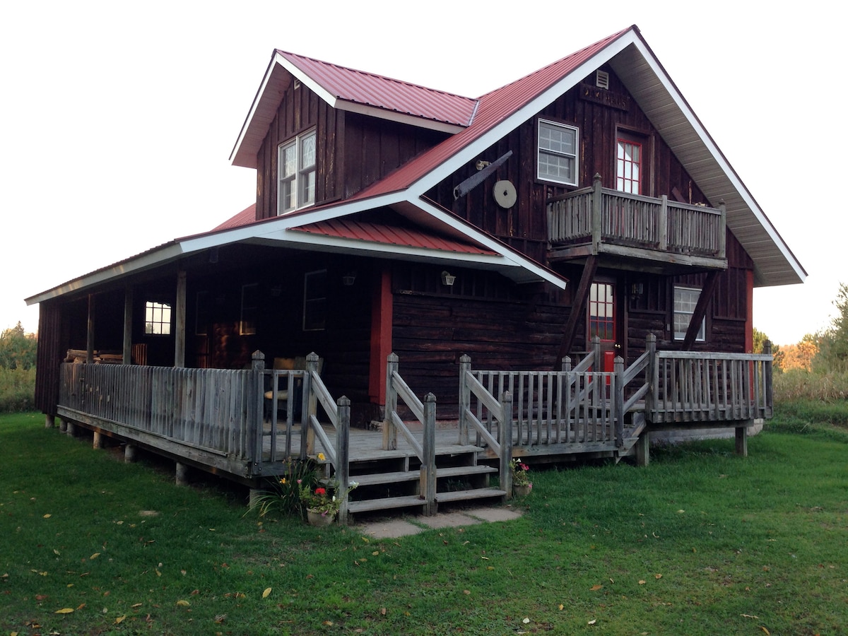 A rustic red-roofed log cabin is showcased with a wrap-around porch and wooden railing. The cabin features two stories, with multiple windows and a balcony on the upper level. The grassy area surrounding the cabin is accentuated by floral plantings.