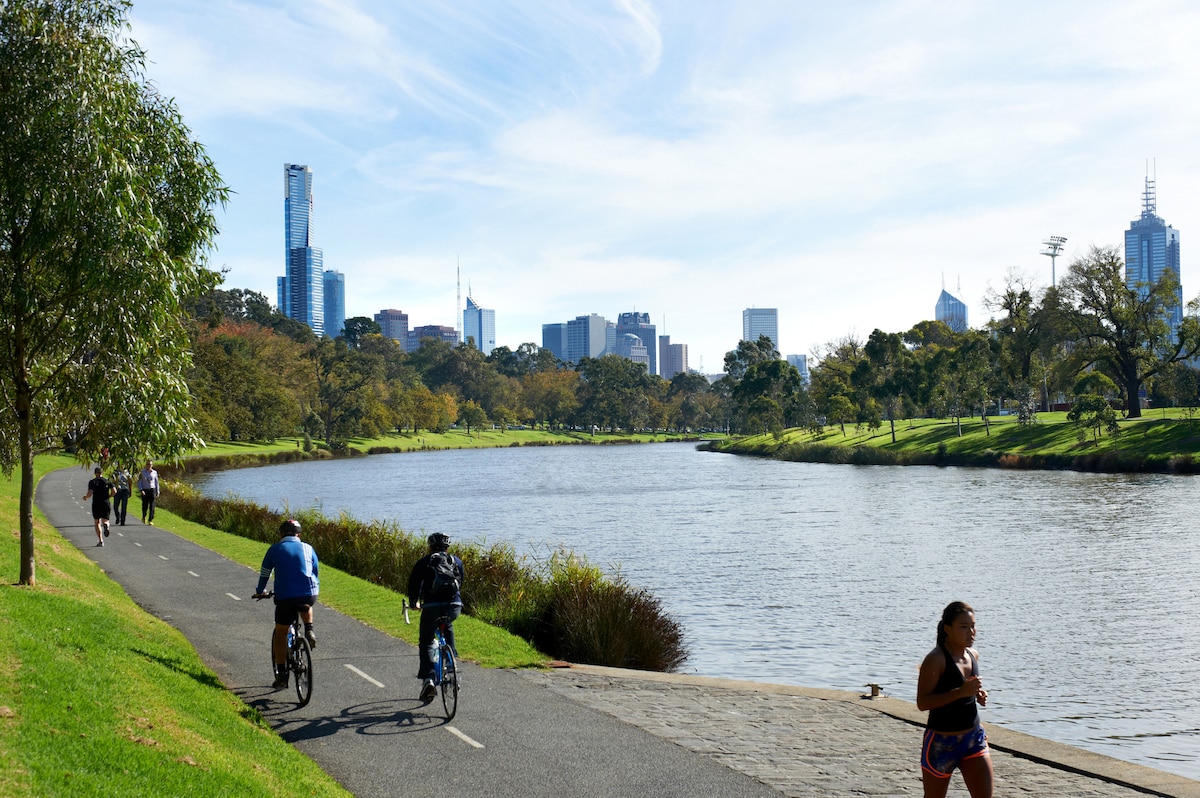 A scenic riverside pathway features cyclists and joggers enjoying the outdoors. Lush greenery lines the riverbank, while the Melbourne skyline is visible in the background under a clear blue sky, creating a pleasant recreational atmosphere.