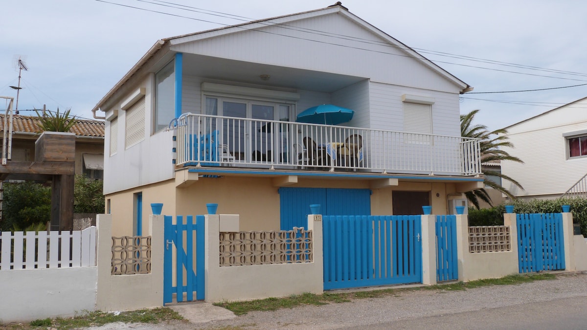 The exterior of a two-story chalet is displayed, featuring a light color facade with blue accents. A balcony is visible on the upper level, furnished with outdoor seating under a blue umbrella. A white fence surrounds the property, providing a welcoming feel.