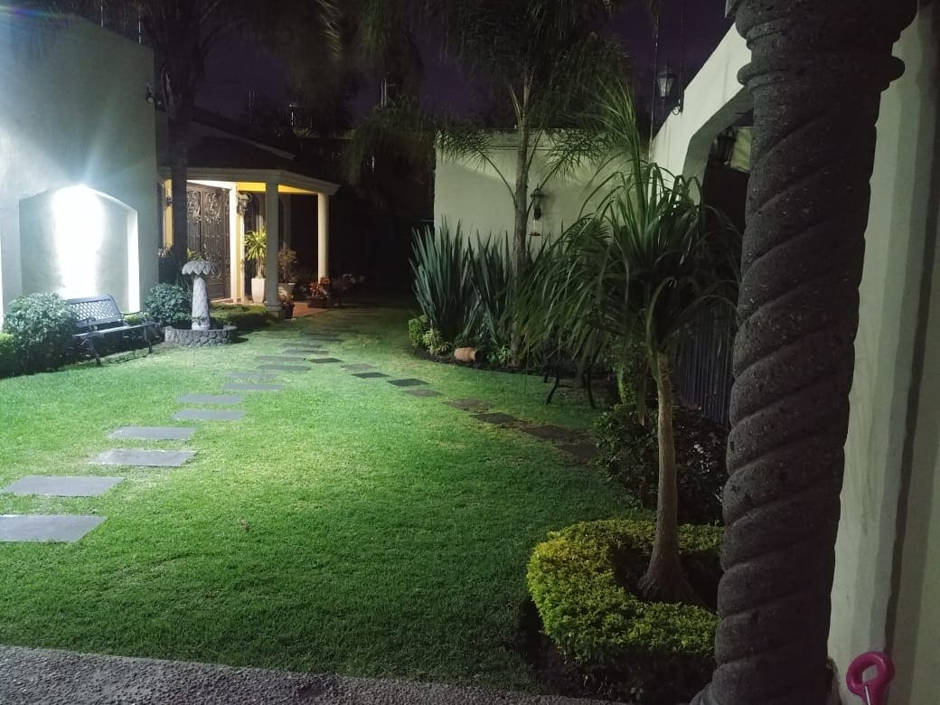 A well-maintained garden features neatly trimmed grass and a path lined with stepping stones. Lush plants and decorative shrubs are present, accompanied by a statue and outdoor seating. Soft lighting illuminates the area, creating a serene atmosphere suitable for relaxation.