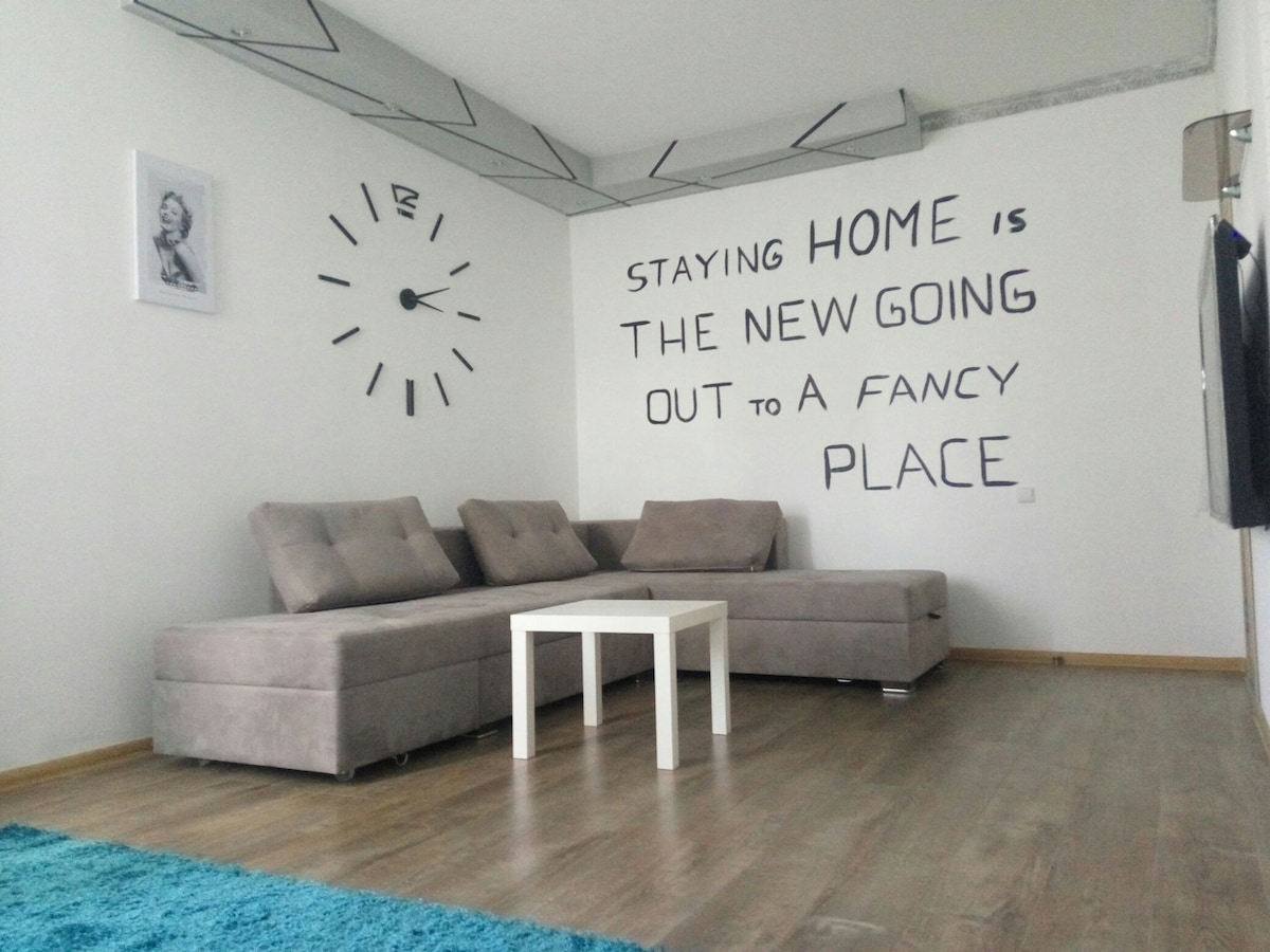 A modern living space is showcased, featuring a sectional sofa in a light gray tone positioned next to a small white coffee table. A bold wall art message is displayed, paired with a wall clock, while natural light illuminates the room's clean lines and wooden floor.
