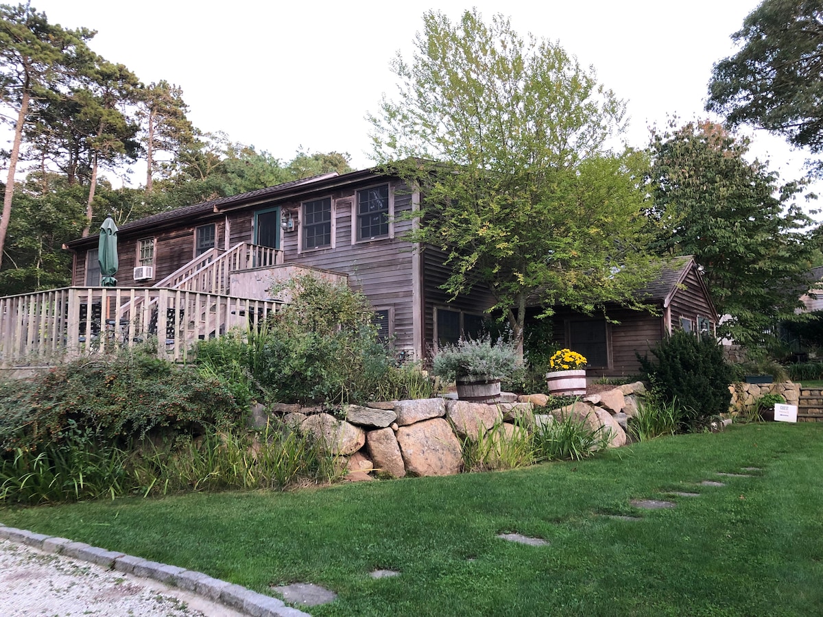 An exterior view of a two-story wooden home surrounded by lush greenery and landscaping. A spacious deck with a green umbrella leads to the entrance, while large stone accents border the grassy yard. The natural setting offers a sense of privacy.