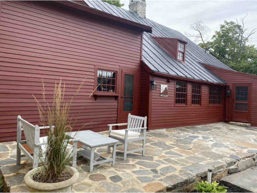 A red wooden exterior of the antique home features a stone patio area. Two white wooden chairs and a small table are positioned on the patio. The landscape includes tall grass in a planter, and windows with grilles provide a glimpse into the cozy interior.