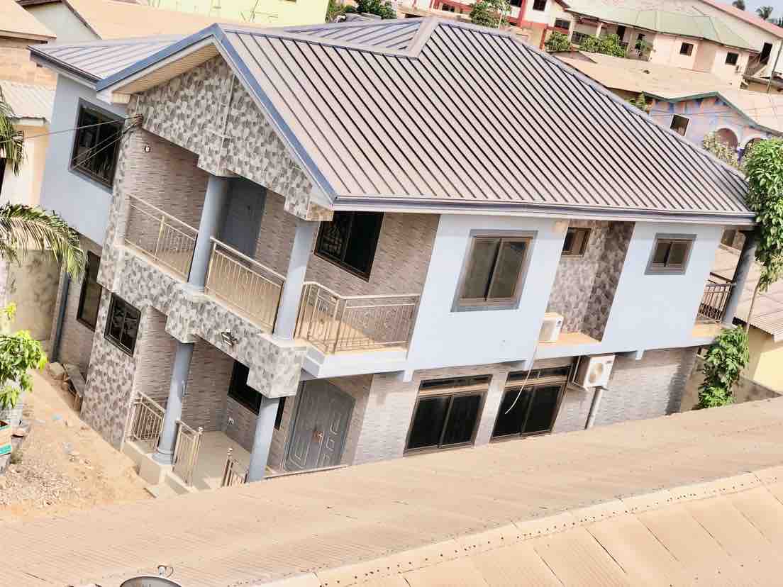 A two-story building is depicted with a modern exterior design featuring a mix of stone and painted surfaces. Balconies with decorative railings are visible, and large windows allow for natural light. An air conditioning unit is mounted on the ground floor.