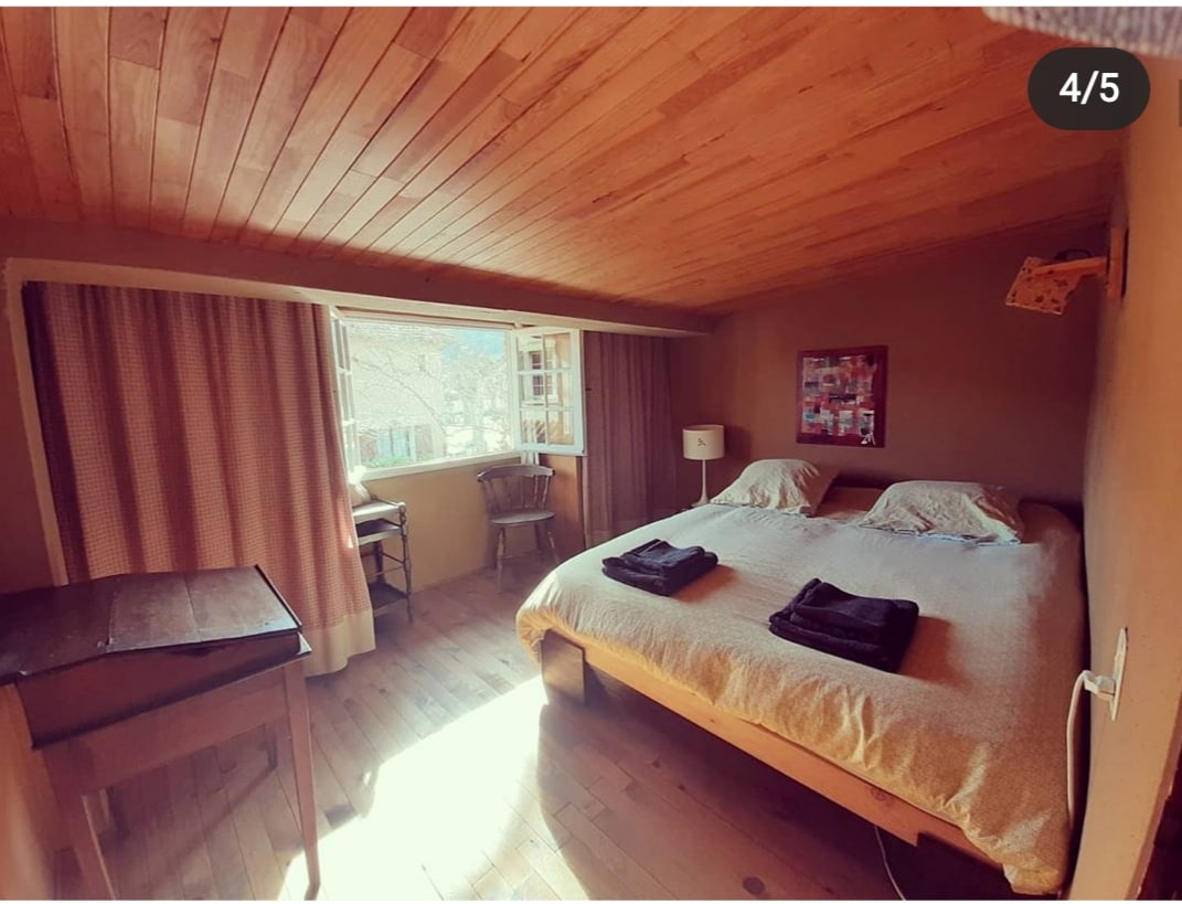 The bedroom features a spacious bed with neatly arranged dark towels on top. A wooden table and a chair are present, alongside a window allowing natural light to illuminate the room, complemented by light brown wooden paneling and soft curtains.