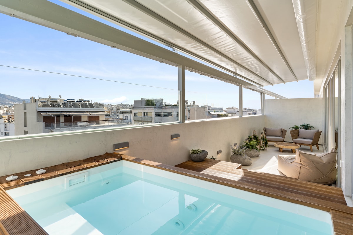 A private balcony showcases a heated plunge pool surrounded by wooden decking. Comfortable seating areas are positioned beside potted plants, offering a relaxed space with panoramic views of the city skyline under clear blue skies. Natural light fills the area through expansive sliding walls.