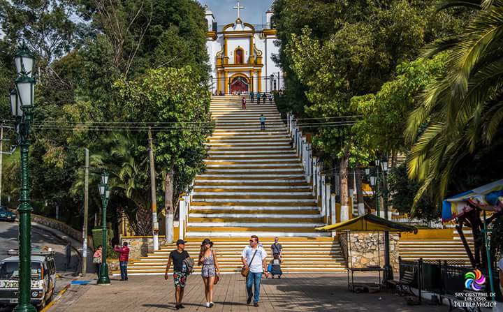 A vibrant scene captures visitors walking up the wide steps towards a prominently featured church, flanked by lush greenery. The structure, adorned with a cross and colorful details, stands against a clear sky, creating a welcoming atmosphere for exploration.