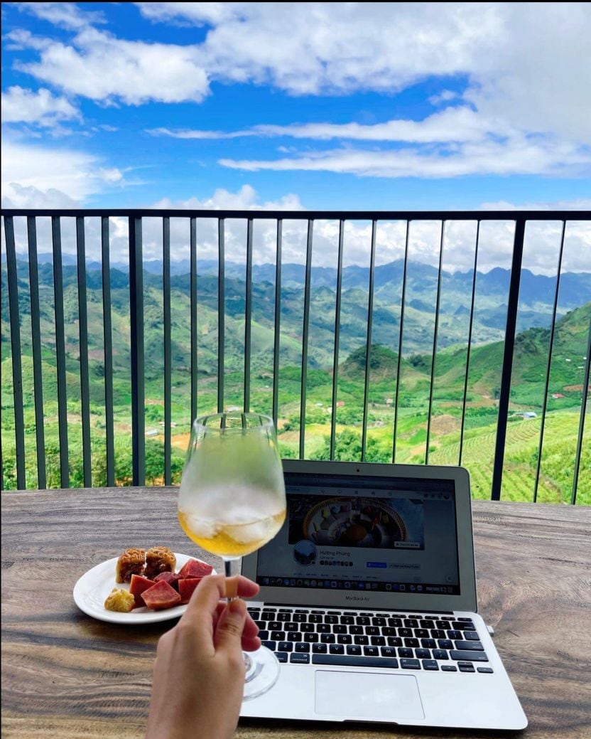A clear view of a mountain landscape unfolds beyond a balcony railing. A laptop rests on a wooden table, adorned with a plate of assorted fruits and bites. A hand holding a glass of beverage is positioned gracefully, enhancing the serene atmosphere.