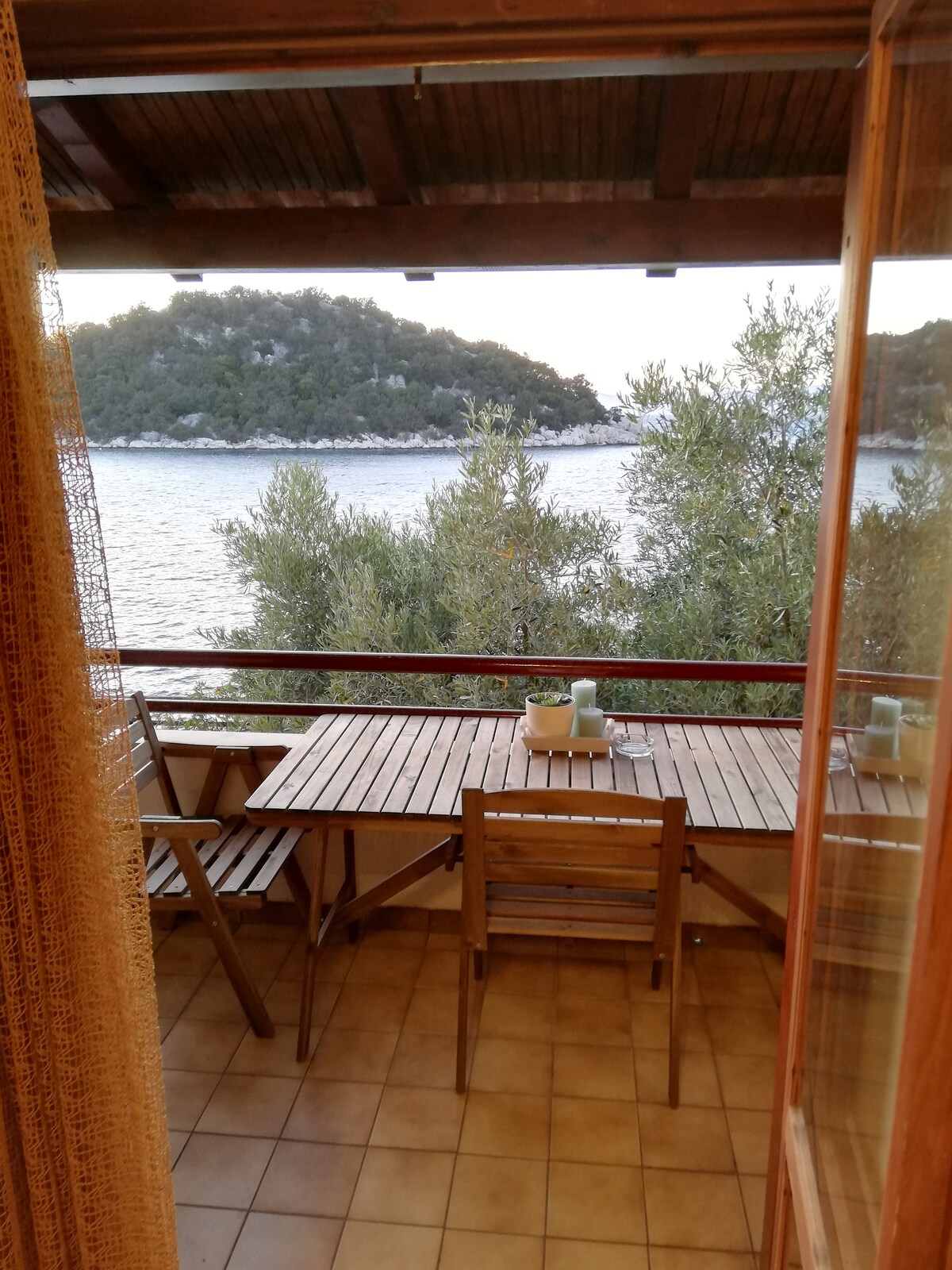 A sunlit balcony is seen, featuring a wooden table with two chairs positioned beside it. The serene waterfront landscape is visible, framed by lush greenery and gentle waves. The scene invites relaxation, with a calm atmosphere created by the natural surroundings.