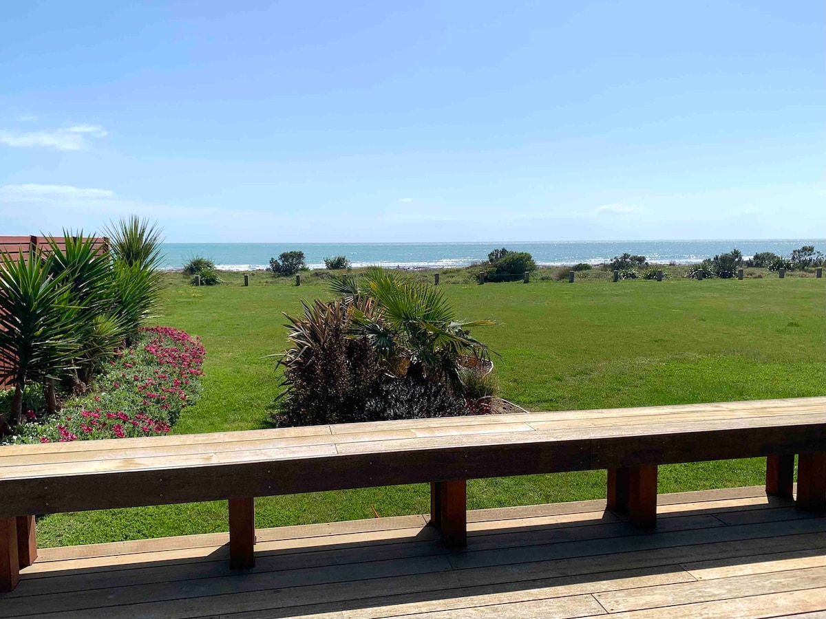 A spacious outdoor deck provides unobstructed views of the ocean and lush green landscape. The foreground features a wooden railing along the deck, while tropical plants and colorful flowers create a vibrant border leading to the serene beachfront beyond.
