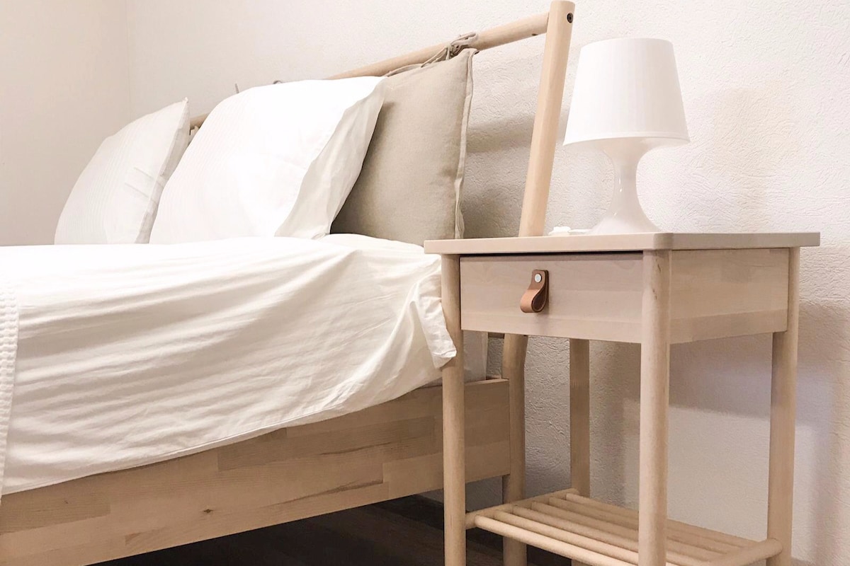 A minimalist bedroom is presented, featuring a wooden bed with a crisp white duvet and beige throw pillows. A small nightstand with an open shelf is positioned beside the bed, topped with a simple white lamp, enhancing the room's clean design.