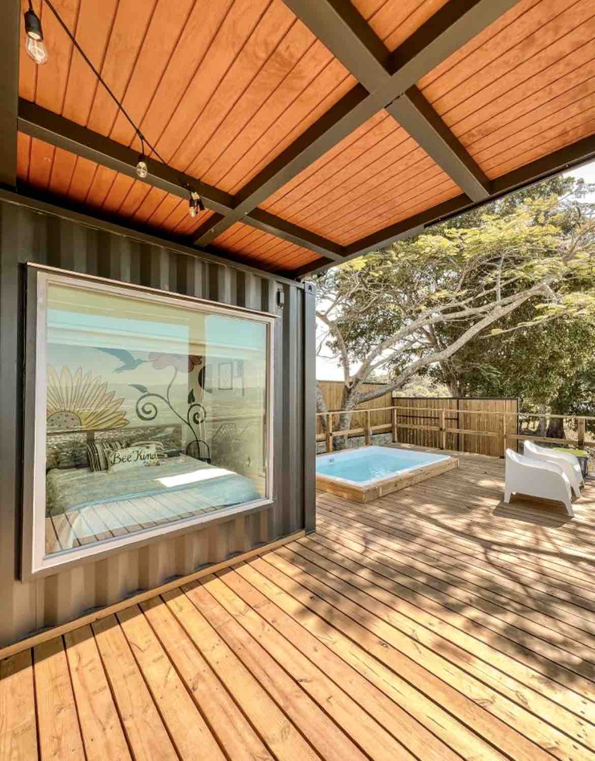 An outdoor deck features a cozy seating area with a white chair and a small hot tub. A large window reveals a sleeping area decorated with a colorful mural and a spacious bed. Surrounding greenery offers a serene backdrop, enhancing the outdoor retreat.