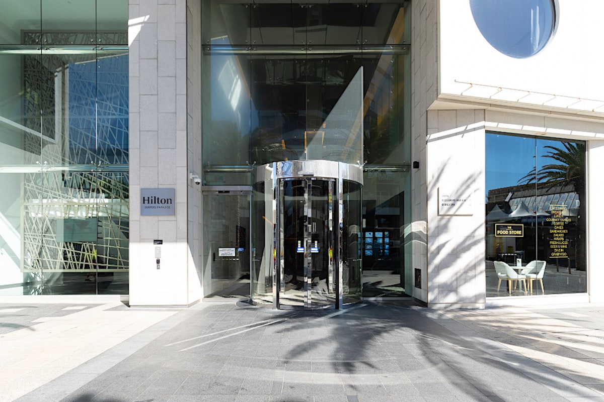 The entrance of the Hilton hotel is showcased, featuring a sleek glass revolving door. The outdoor area is paved with gray tiles and palm trees are visible nearby, contributing to a modern and welcoming atmosphere.