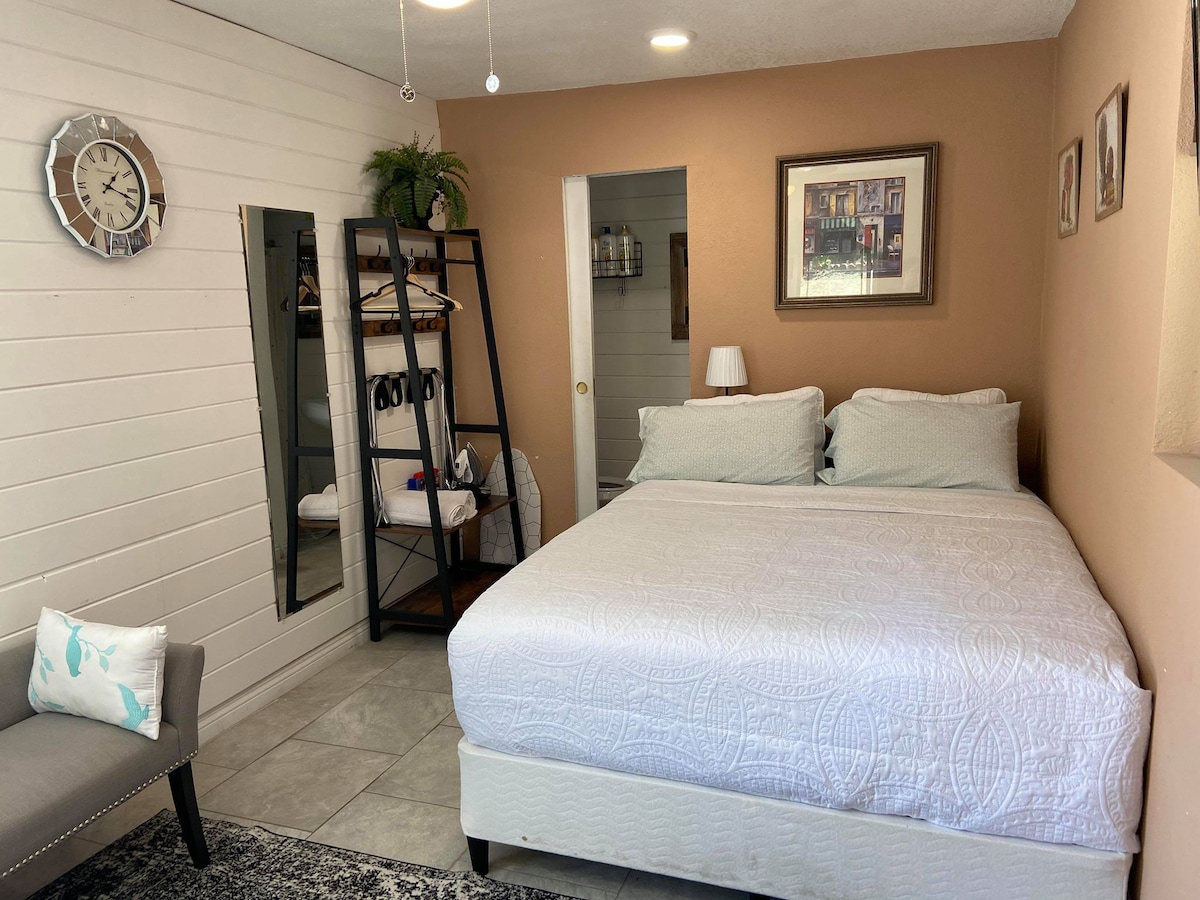 A bright studio space features a queen bed with soft bedding, complemented by a bedside lamp. A stylish chair sits nearby, with a mirror and wall decor enhancing the room's functionality. Light tiles and neutral tones contribute to a serene ambiance.