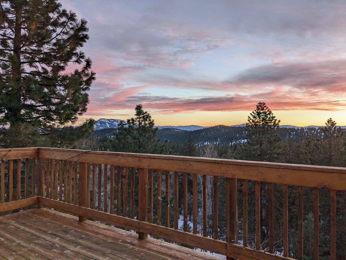 A spacious wooden deck offers panoramic views of the surrounding mountains and forest, bathed in warm hues of sunset. Pines frame the view, emphasizing the serene setting with a tranquil atmosphere. Snow covers the ground beneath the trees, hinting at the winter season.