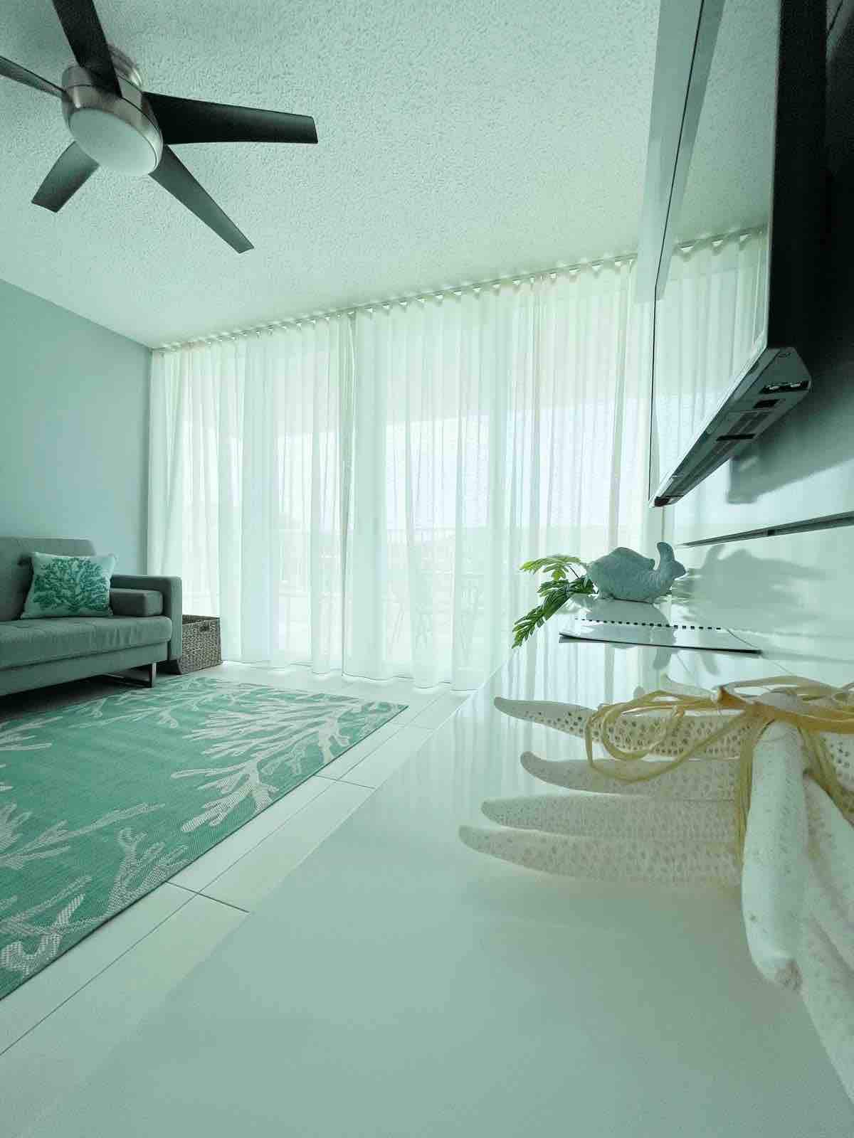 A bright living area showcases a comfortable couch adorned with decorative pillows. Sheer curtains allow soft natural light to fill the space, reflecting off the sleek, light-colored surfaces. A coral-themed area rug provides an inviting touch, complemented by decorative accents and a fan overhead.