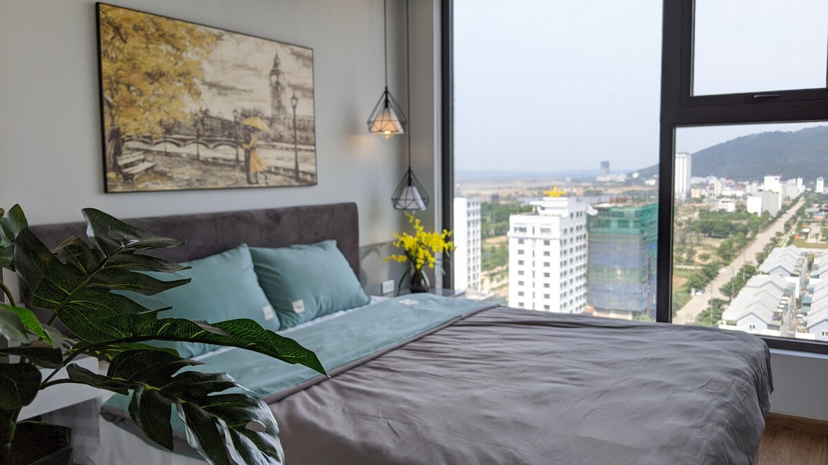 A spacious bed is centered in the room, adorned with soft green and gray linens. A large window offers a panoramic view of the cityscape, while a small potted plant and yellow flowers add a touch of nature to the bright interior.