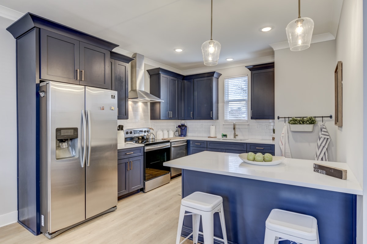 The kitchen features navy blue cabinetry and stainless steel appliances, including a refrigerator and oven. A spacious island is complemented by three white stools, and natural light illuminates the area through a nearby window. Fresh green apples sit on the counter for a pop of color.