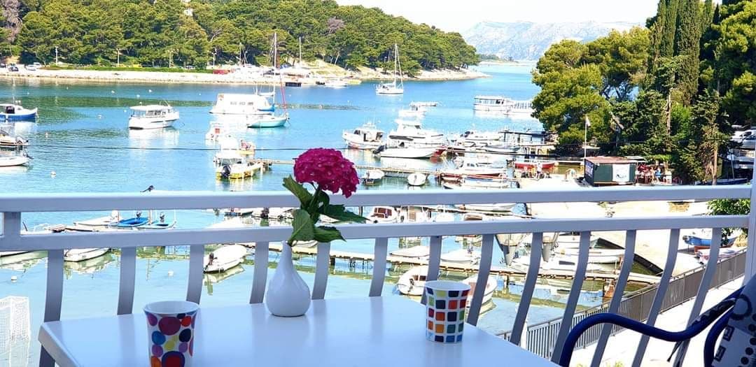 A balcony view overlooks a marina filled with boats, surrounded by lush greenery and distant hills. A white table is set with a small vase holding a flower and two colorful cups, providing an inviting space to enjoy the scenery.