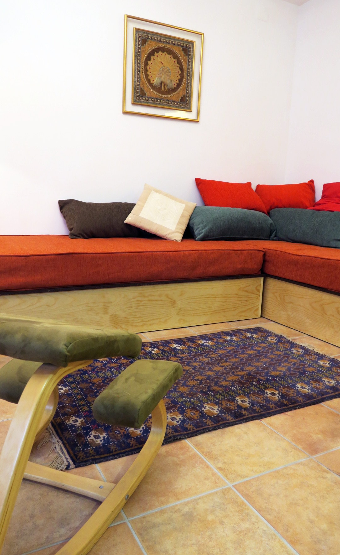 A cozy seating area features a low wooden bench with colorful cushions in red and green tones. A small green rocking chair is placed nearby on top of a decorative rug, enhancing the inviting look of the space.