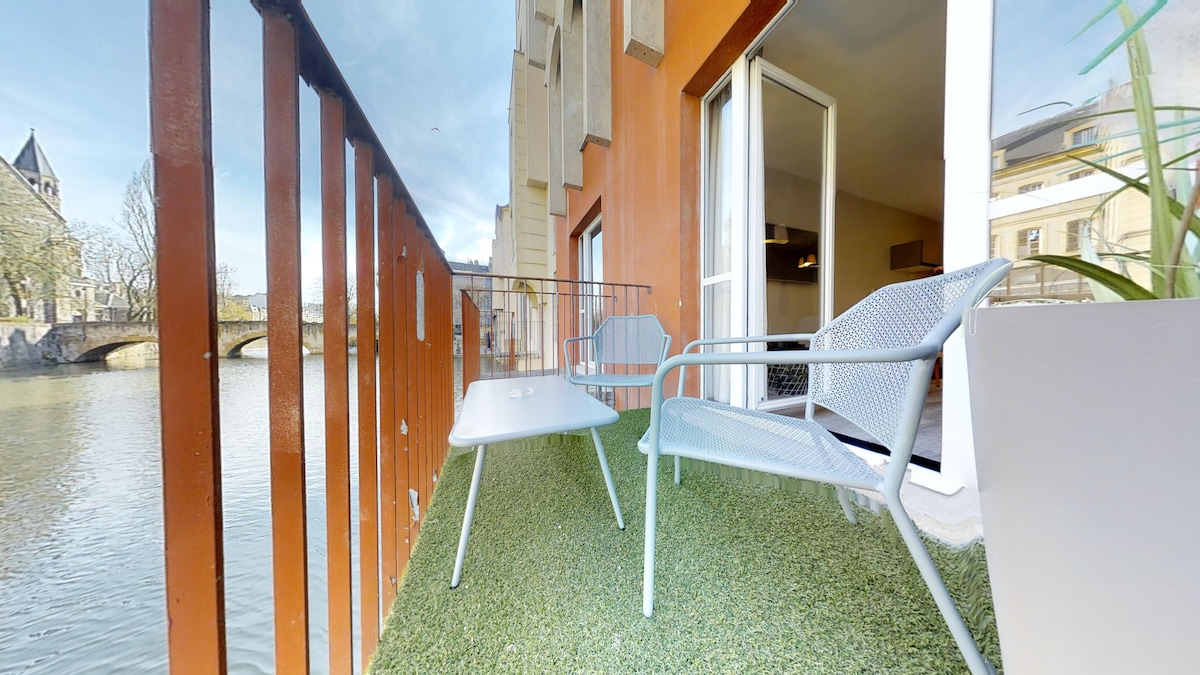 A private terrace offers views of the Moselle River, furnished with two light-colored chairs and a small table. The space is bordered by a railing and features a faux grass surface, creating a relaxing outdoor environment.