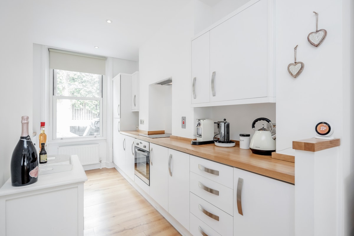 A modern open-plan kitchen is displayed with sleek white cabinets and wooden countertops. The kitchen features a built-in oven, kettle, toaster, and various appliances. Bright natural light enters through a large window, enhancing the clean and functional design.