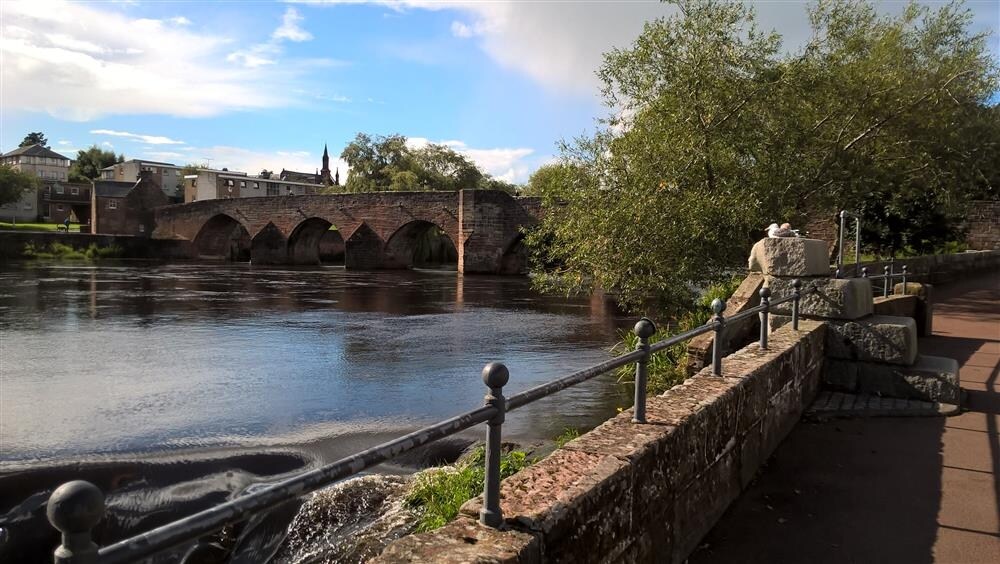 A scenic riverside view features an ancient stone bridge spanning the flowing water, with arches visible under the clear sky. Lush greenery lines the bank, and a pathway runs alongside the river, offering a tranquil atmosphere for leisurely walks.