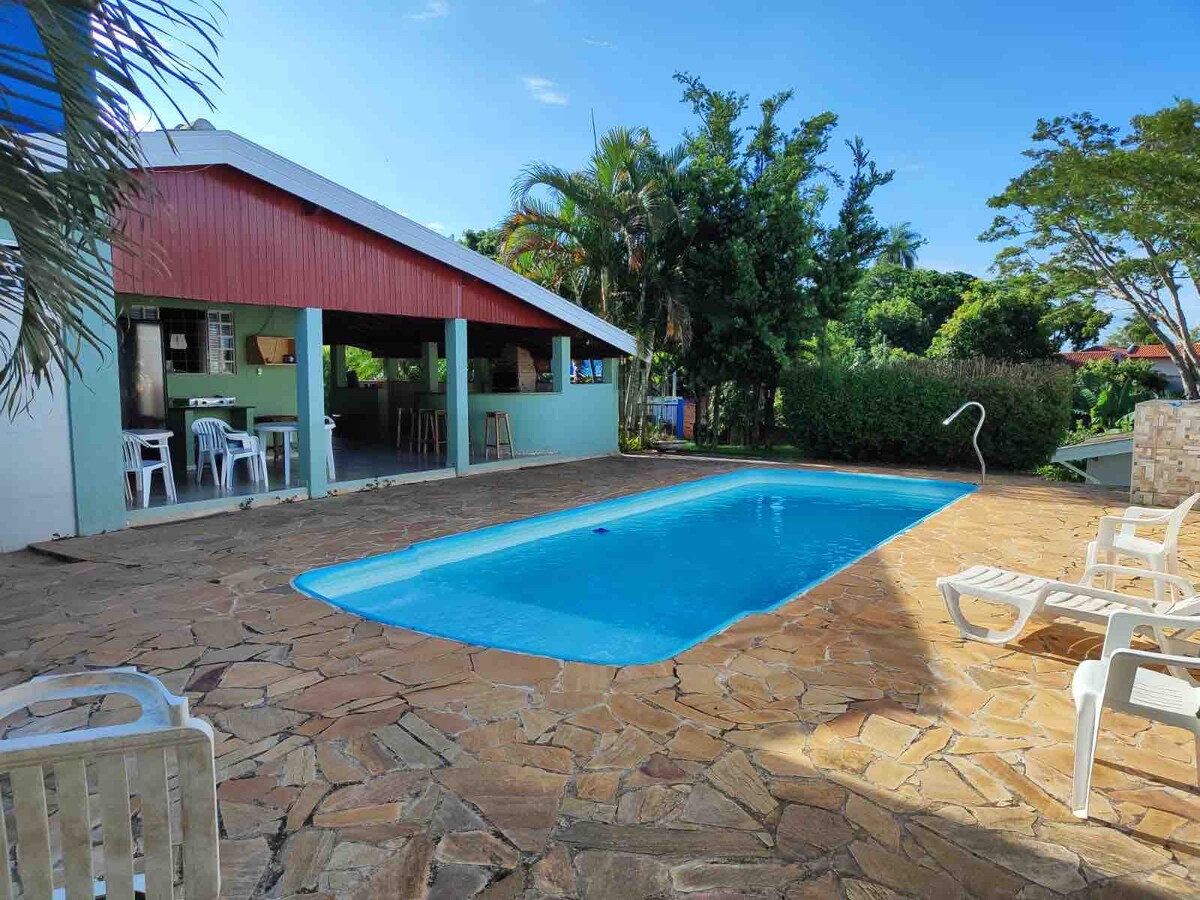 A tranquil outdoor area features a clear blue swimming pool surrounded by a spacious stone patio. Sun loungers are positioned nearby, and a covered seating area with white chairs and a table is visible in the background, complemented by lush green trees.