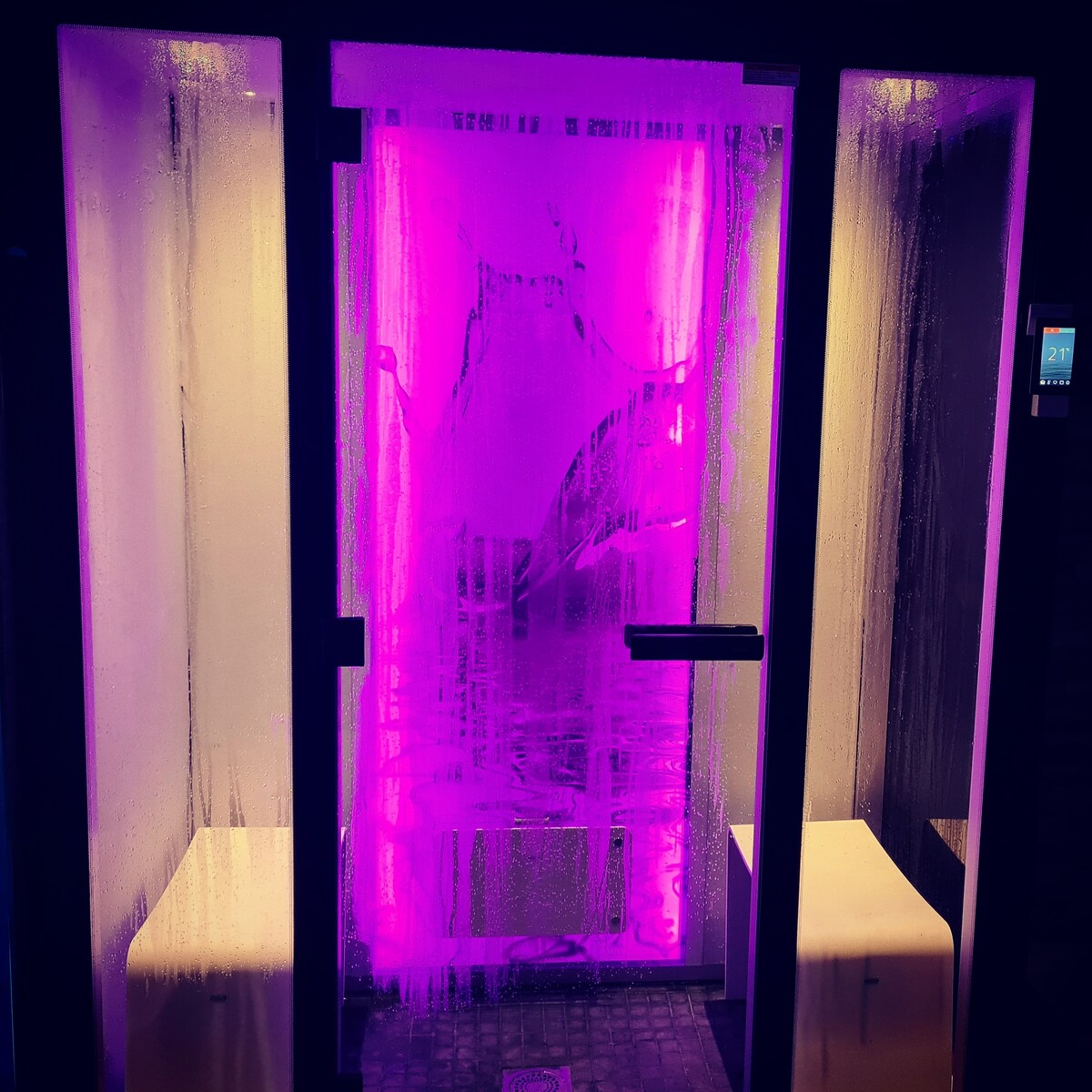 A contemporary steam room features a fogged glass door with vibrant purple lighting. The interior showcases two benches on either side, positioned for relaxation. The warm atmosphere is enhanced by the glow from strategically placed lights, creating an inviting space for rejuvenation.