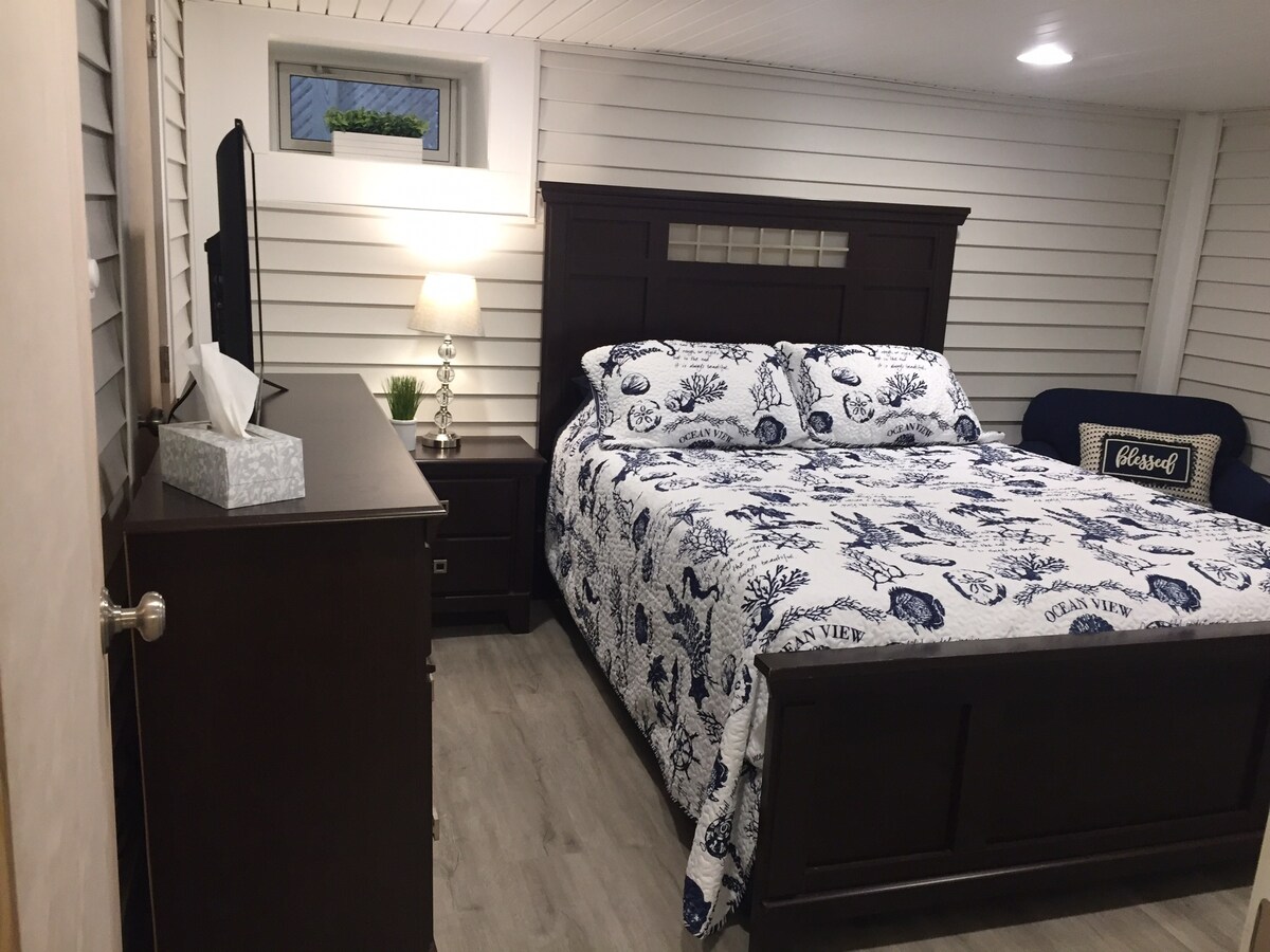 A cozy bedroom suite is featured with a spacious bed dressed in a blue and white patterned quilt. A dresser and nightstand accompany the bed, providing storage. Natural light enters through a small window, and a decorative plant enhances the ambiance.