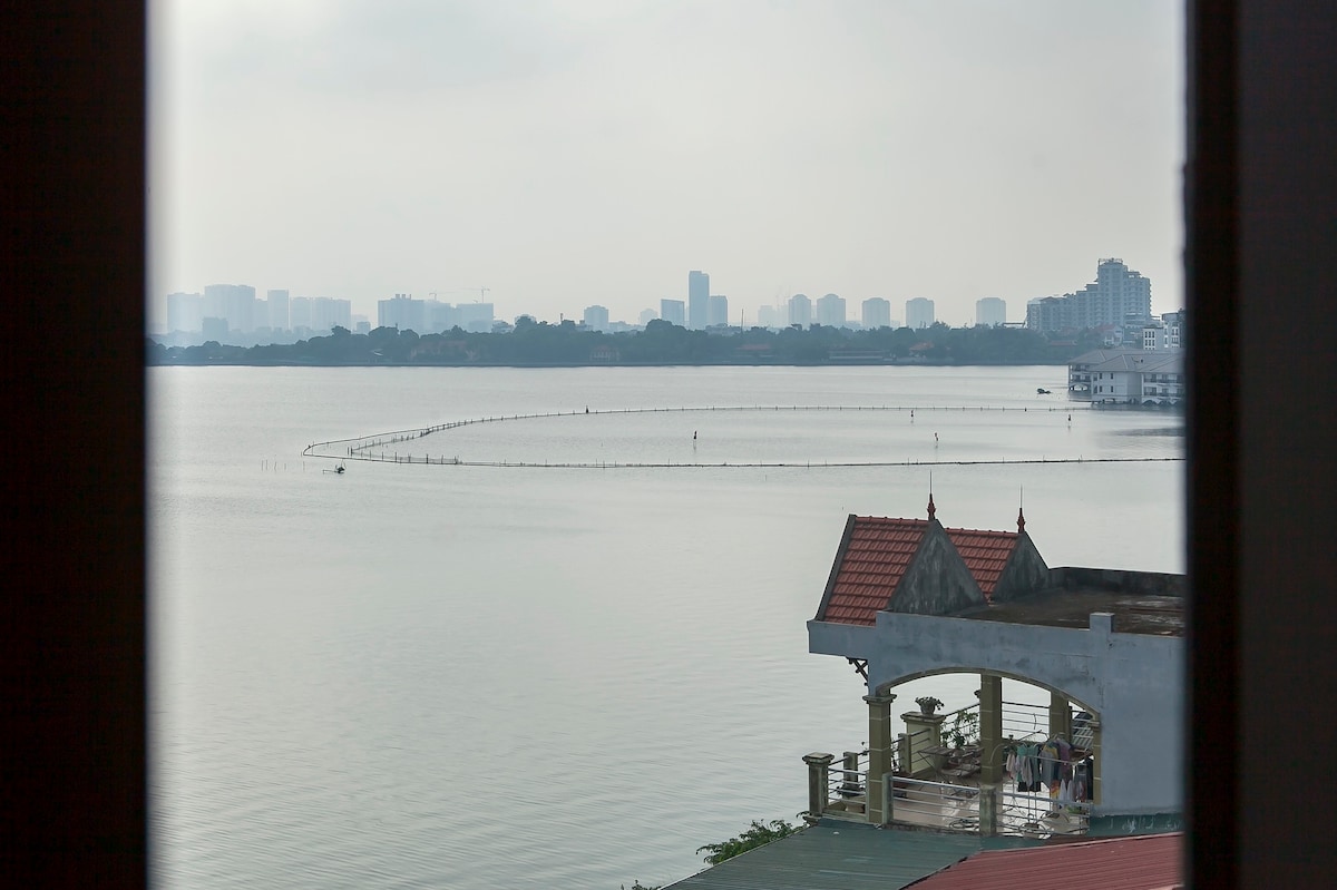 A serene lakeside view is captured through a window, revealing a calm body of water with a circular fishing net visible in the distance. Urban buildings rise along the horizon, while a traditional pavilion with a red-tiled roof is seen in the foreground.