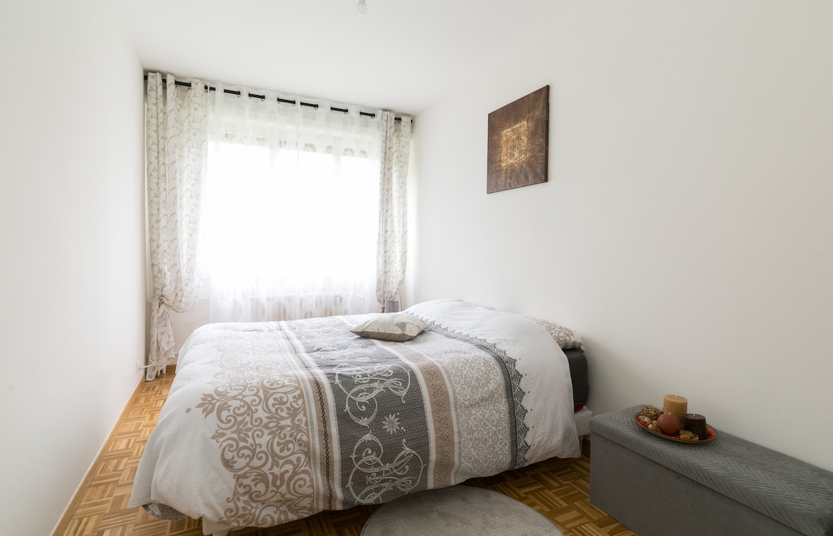 A bright bedroom features a spacious bed adorned with a decorative blanket and a neutral-toned pillow. Natural light filters through sheer curtains, illuminating the room. A small bedside table holds a candle and a decorative tray, enhancing the simple elegance of the space.