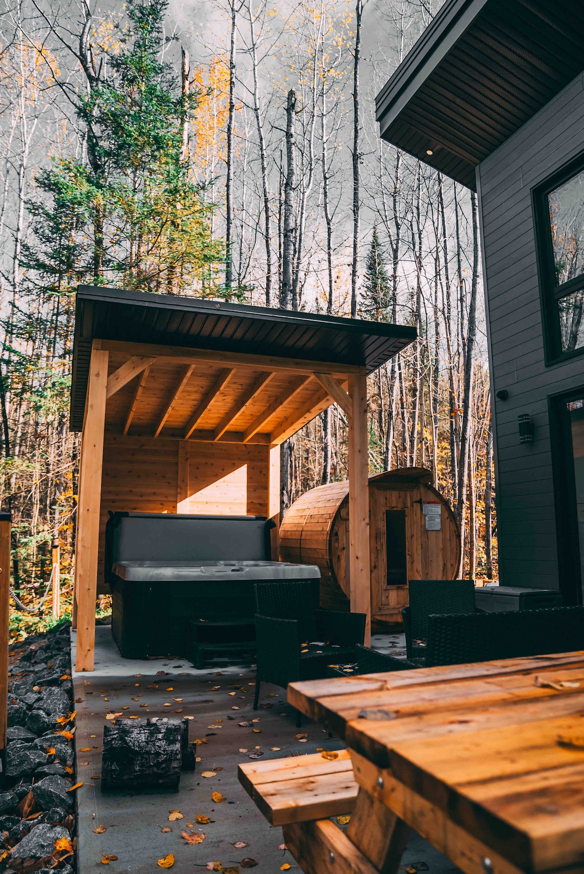 An outdoor spa area is nestled among trees, featuring a hot tub under a wooden canopy. Nearby, a traditional sauna stands ready for use. A wooden dining table is positioned nearby, surrounded by dark wicker chairs, all set on a patio with scattered autumn leaves.
