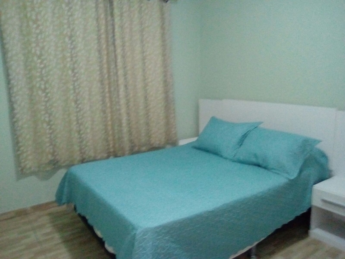 This sleeping area features a spacious bed dressed in a light blue coverlet and matching pillows. Soft, neutral walls complement the light-colored furniture. A window dressed with patterned curtains filters natural light into the room, creating a calm environment.