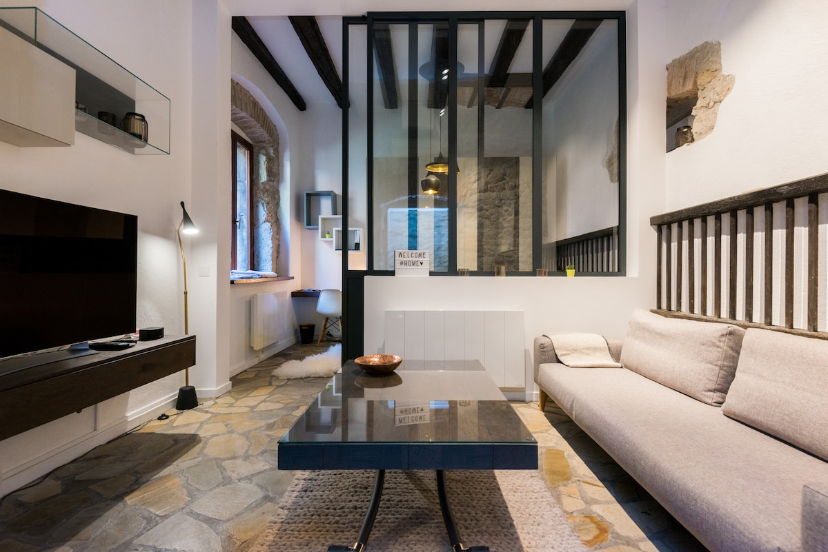 A living area features a modern design with a spacious sofa facing a flat-screen television. Large glass windows allow natural light to fill the room, and a stone floor adds texture. Decorative elements include a minimalist side table and a wall with wooden accents.