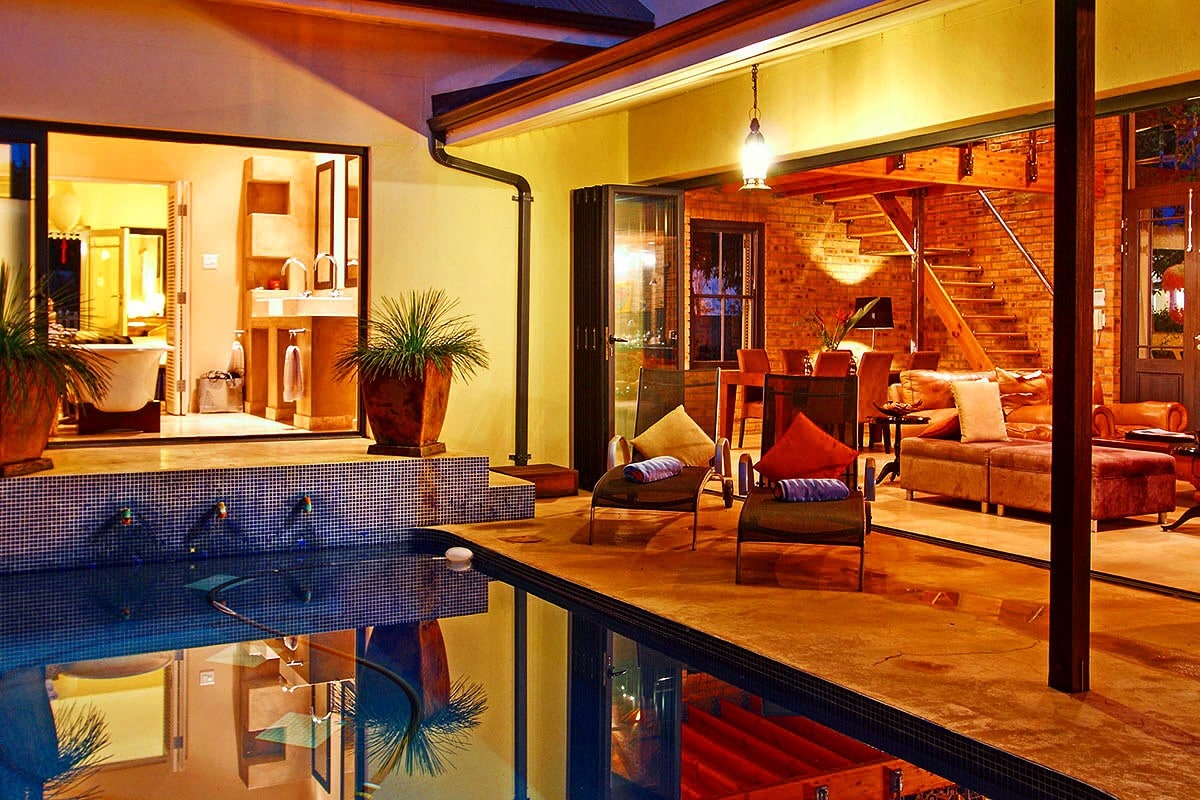 An outdoor area showcases a private pool reflecting the surrounding space. Comfortable seating arrangements, adorned with cushions, are positioned nearby. Interior spaces are visible, featuring warm lighting that highlights a dining area and a staircase, creating a seamless flow between indoor and outdoor living.