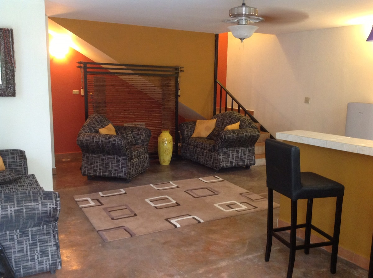 A living area features two patterned armchairs and a coordinating couch, all arranged around a central area rug. A bar-height chair sits at the counter, while a modern ceiling fan provides airflow. Warm lighting highlights the vibrant orange and yellow walls.