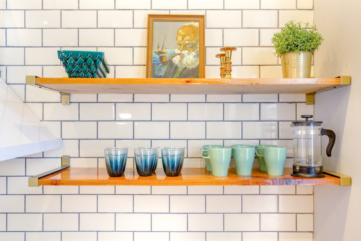 Two wooden shelves are mounted against a white subway tile backdrop. A variety of glassware and ceramics are displayed, including six blue glasses and several green mugs. A small plant in a gold pot and a framed painting are positioned on the upper shelf.