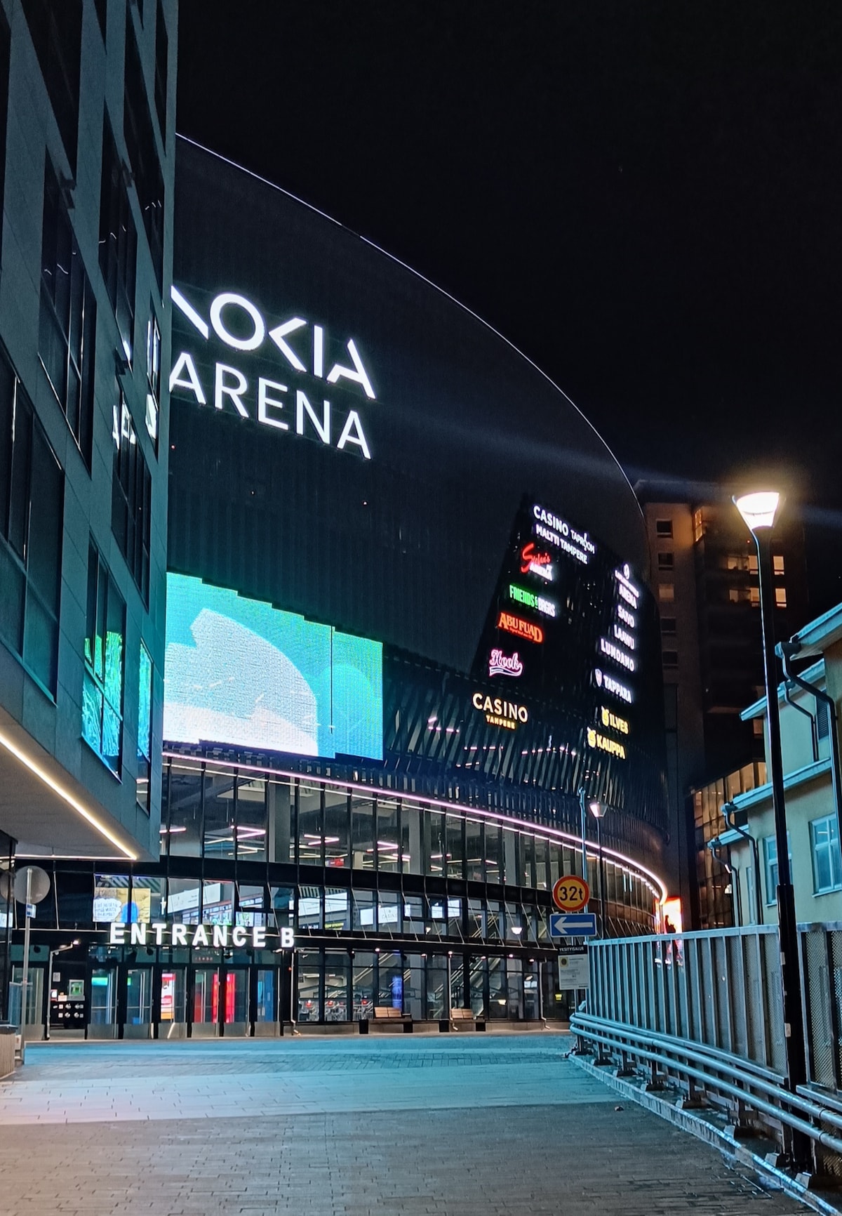A night view of Nokia Arena's exterior, showcasing its illuminated signage. The entrance is clearly marked with 'ENTRANCE B,' while the sleek, modern architecture reflects the urban setting. Streetlights enhance visibility along the sidewalk leading to the arena.