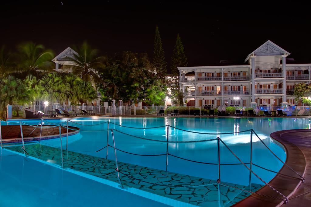 The expansive infinity pool is illuminated at night, reflecting soft blue tones. Lounge chairs line the edges, surrounded by lush greenery. The colonial-style residence is visible in the background, showcasing multiple balconies. Soft lighting highlights the pathways and palm trees in the serene environment.