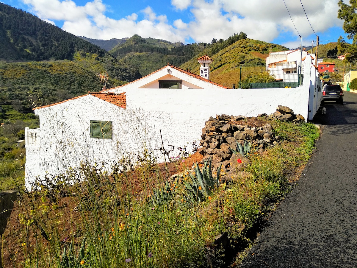 A scenic view of a white-walled house nestled among rolling green hills. The surrounding landscape features vibrant vegetation and rocky outcrops, with distant mountains visible under a partly cloudy sky. A narrow road leads up to the property, enhancing its rural appeal.