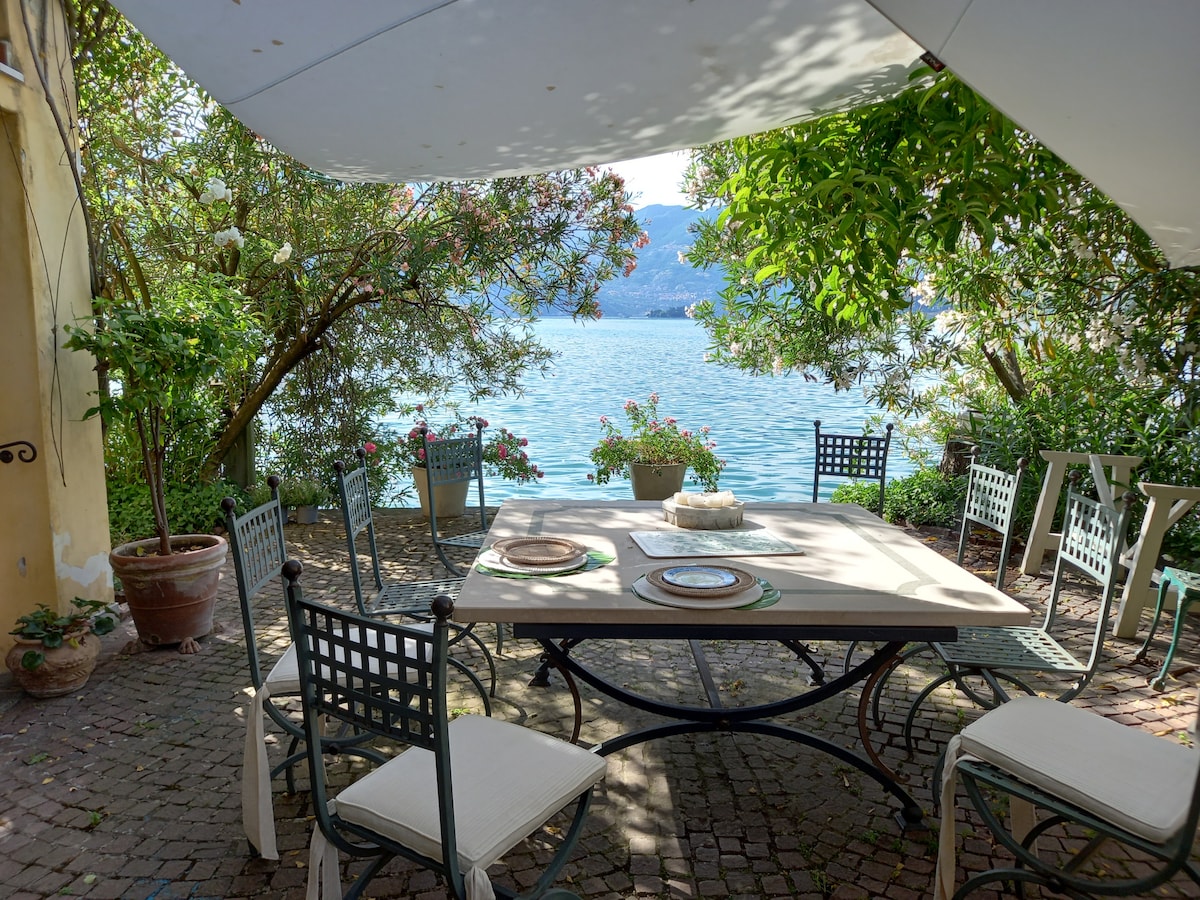 A shaded outdoor dining area features a large table surrounded by several chairs, with views of the shimmering lake through lush greenery. Potted plants add to the inviting ambiance, creating a serene setting for meals al fresco.