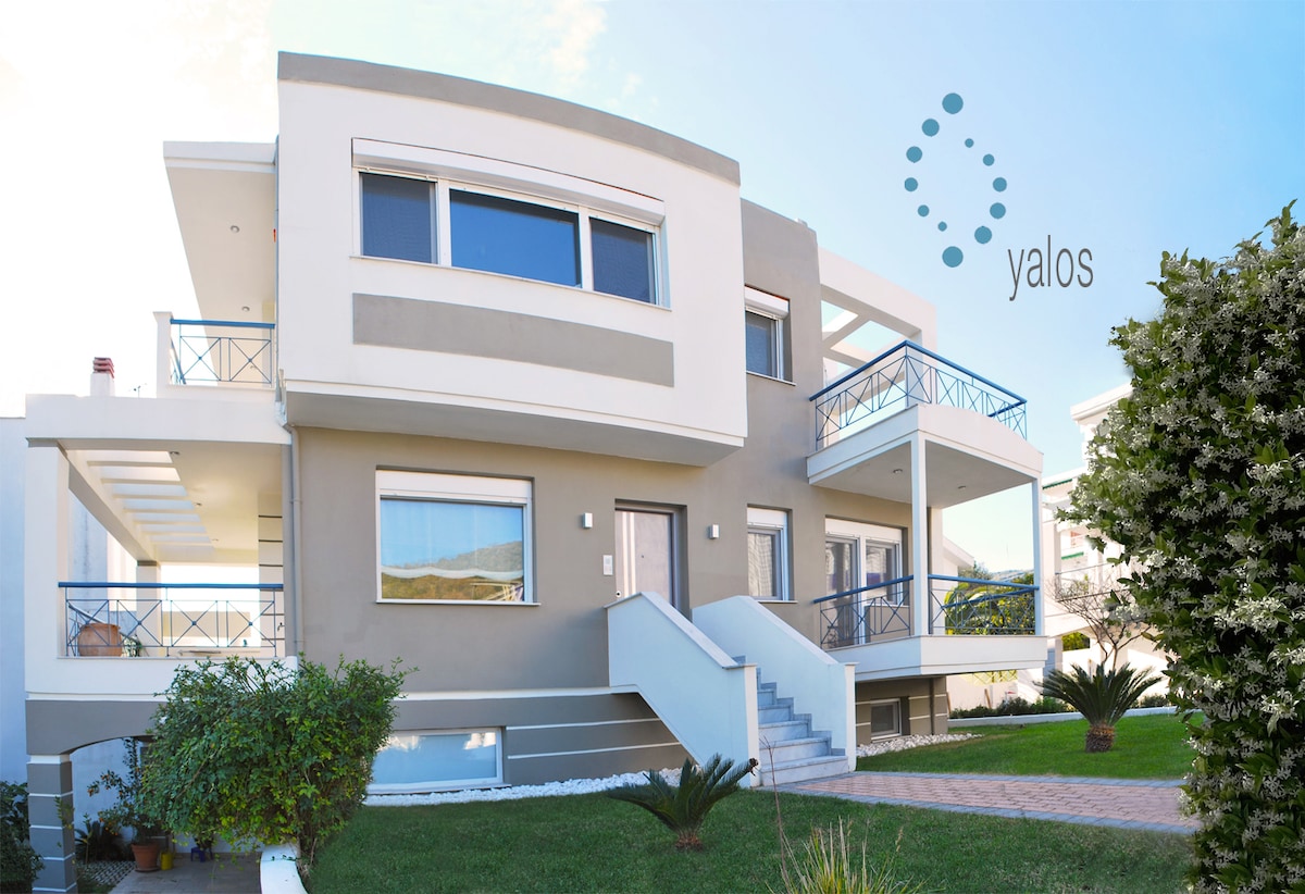 The exterior of a contemporary two-story beach house is shown, featuring clean lines and balconies on both levels. Lush greenery surrounds the property, with well-maintained pathways leading to the entrance. Large windows allow natural light to fill the interiors, enhancing the inviting atmosphere.